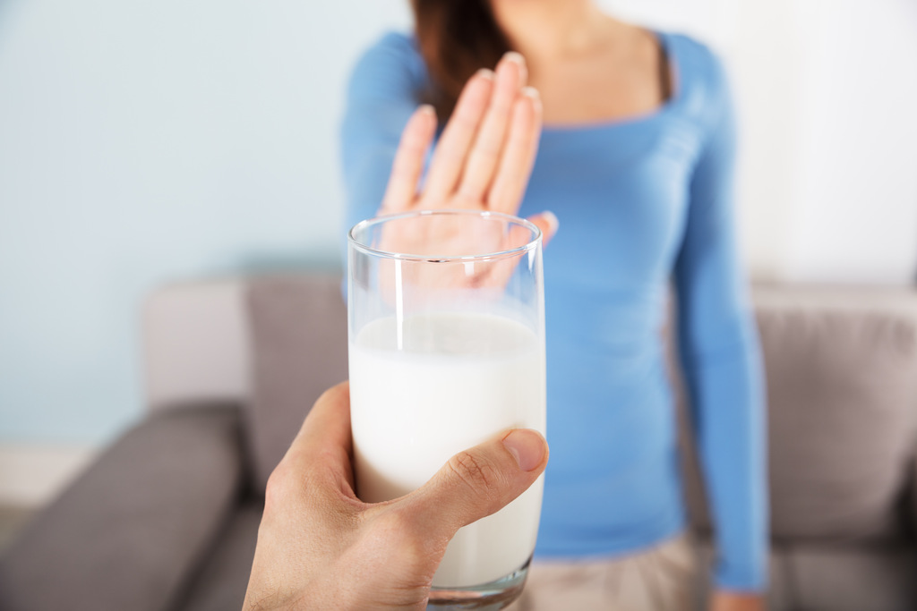 Mulher ao fundo faz um gesto com a mão recusando um copo de leite que é oferecido em primeiro plano. Representa a recusa de laticínios e alimentos a evitar que soltam o intestino.