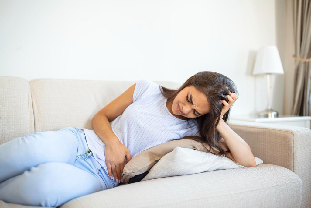 Mulher deitada em um sofá bege com expressão de dor e desconforto, segurando a barriga com uma mão e a cabeça com a outra. A cena representa o mal-estar e as cólicas abdominais, sintomas frequentemente investigados ao buscar pelas principais causas da diarreia amarela.