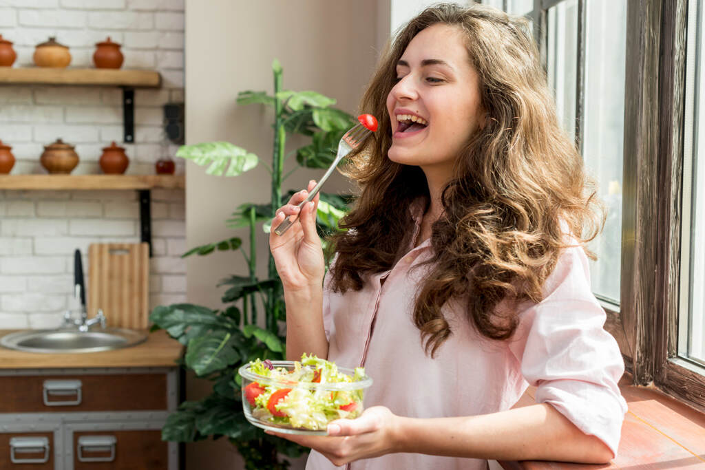 Mulher jovem e sorridente comendo uma salada fresca em uma tigela de vidro na cozinha, ilustrando a alimentação leve como um dos principais cuidados contra a diarreia.