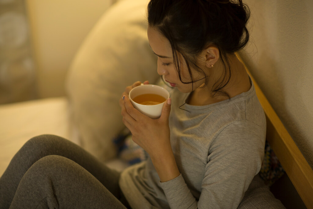 Mulher sentada na cama bebendo uma xícara de chá quente para acalmar o estômago e reduzir o desconforto de uma dor de barriga.